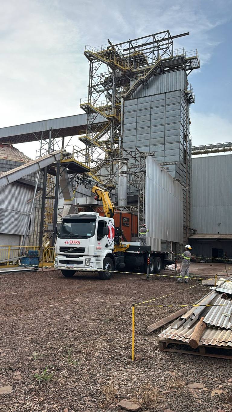 Caminhão munck posicionado em pátio de agroindústria, com estruturas metálicas